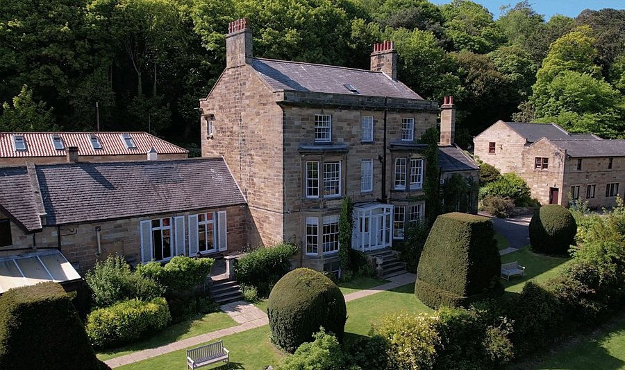 A historic stone building surrounded by manicured gardens and tall hedges, set against a wooded backdrop.