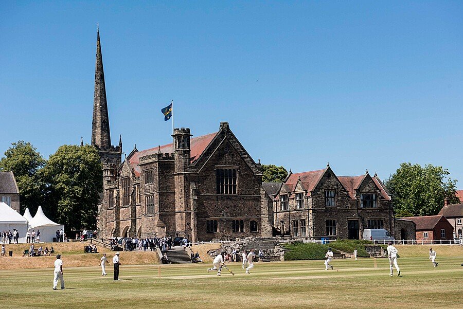 Historic school building with a tall spire and red-tiled roofs, overlooking a cricket match being played on the school grounds.
