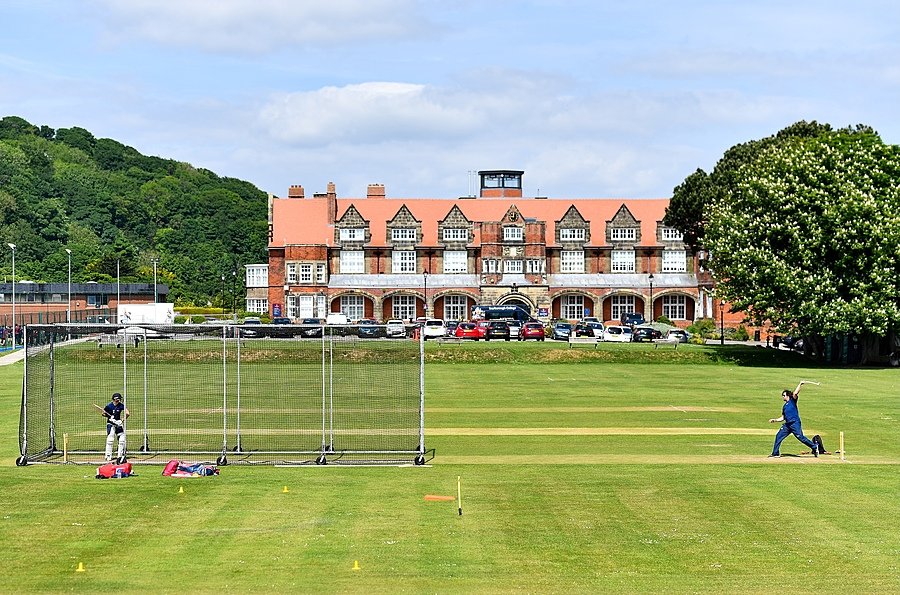 A cricket match is taking place on a large green field in front of a grand red-brick school building with a red-tiled roof and several cars parked outside.