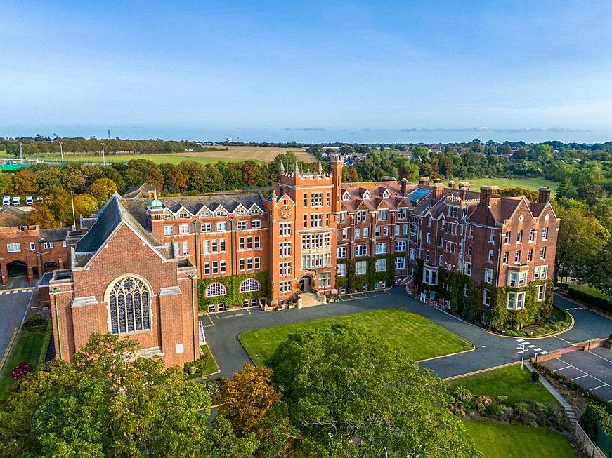 Aerial view of a large red-brick school building with a clock tower, chapel, and green lawns surrounded by trees and open countryside.