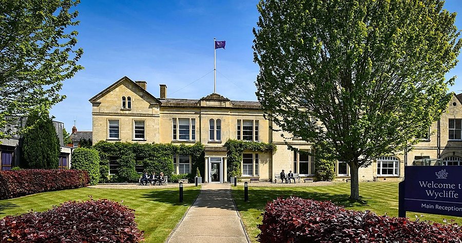 A yellow-brick building with pupils sitting on benches in front of it in the sunshine