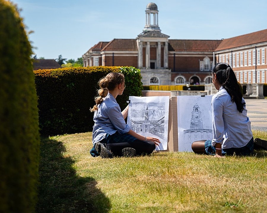 Two pupils sit on the grass sketching the school’s grand clock tower and buildings on a sunny day.