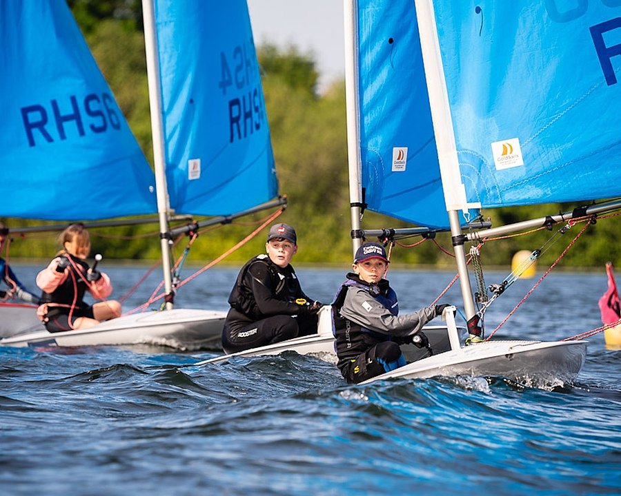 Pupils sailing small boats with bright blue sails on a calm lake, concentrating as they steer and adjust their ropes.