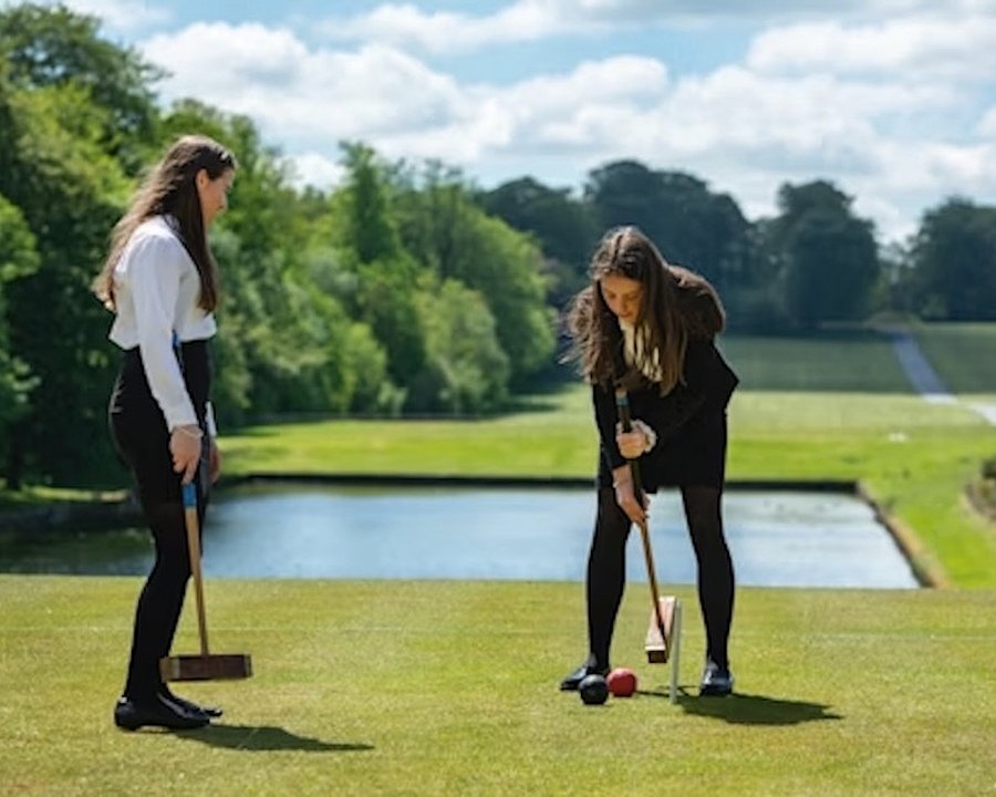 Two students playing croquet on a lawn beside a reflecting pool, surrounded by trees and greenery under a bright sky.