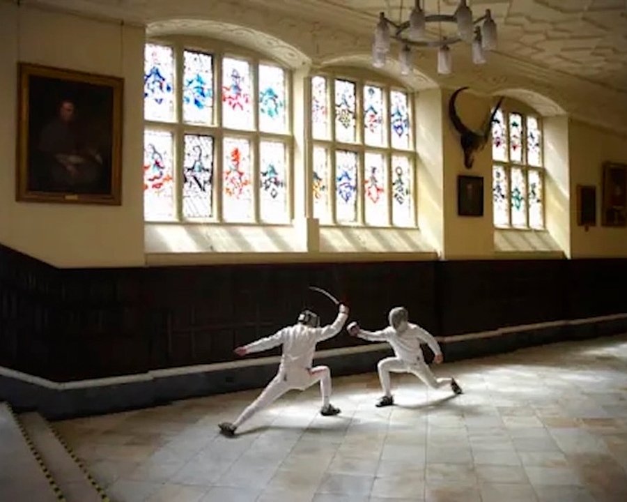 Two fencers in white uniforms sparring inside a historic hall with stained-glass windows and portraits on the walls.