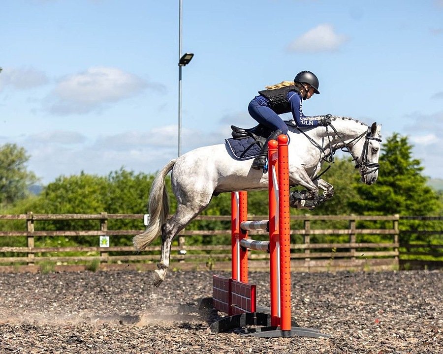 A student riding a white horse during a show jumping session, as the horse clears a red obstacle in an outdoor arena.