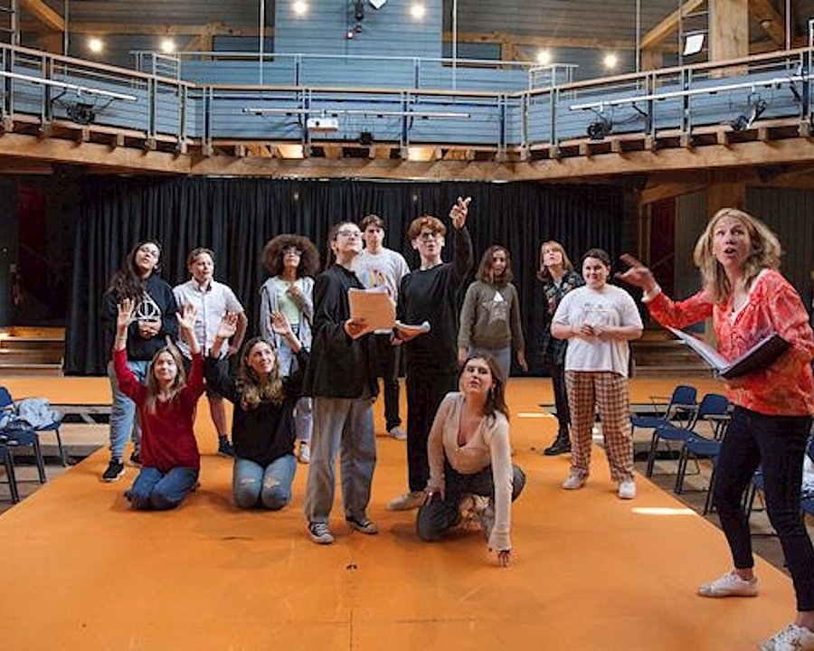 A group of students rehearsing a play on stage in a modern wooden theatre with seating arranged in the round.