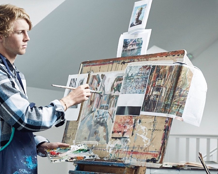 A focused student painting at an easel with another student working in the background in an art classroom.