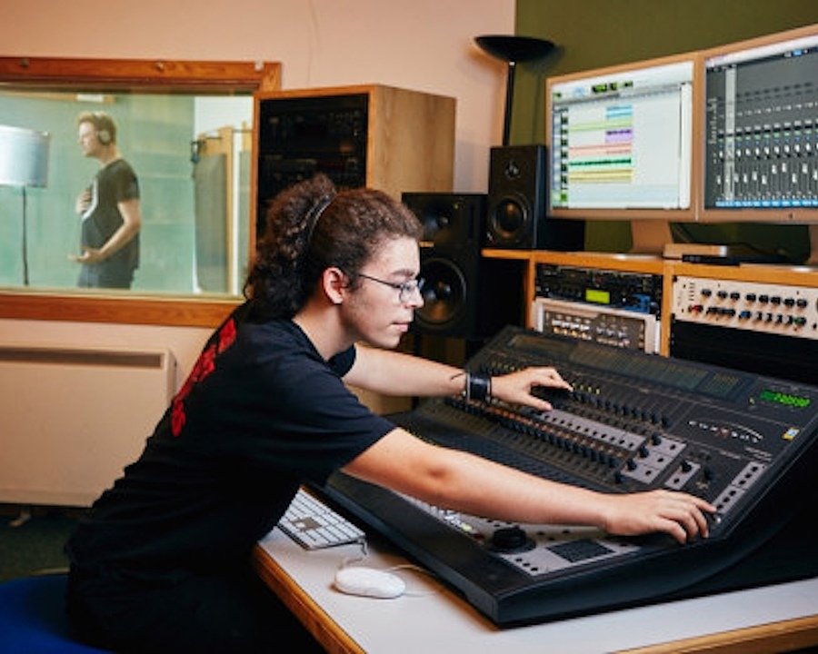A student operating an audio mixing console in a recording studio while another performs in the booth.