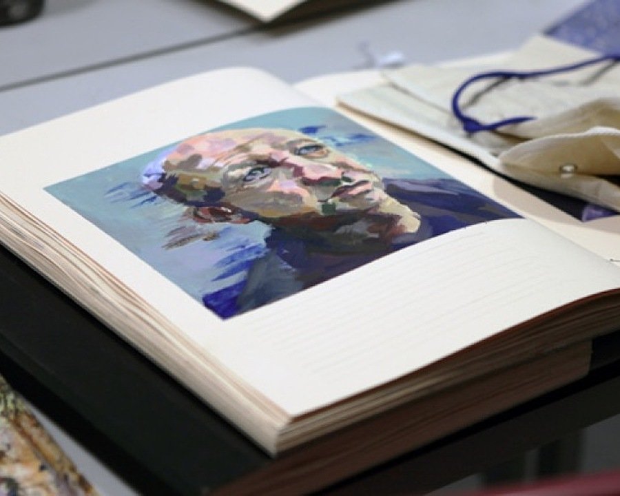 An open sketchbook on a table showing a colourful painted portrait beside art materials.