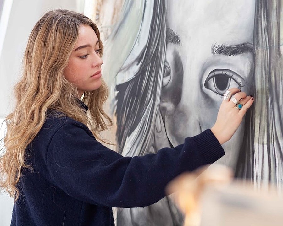 Girl in blue jumper painting a portrait of a girl in front of a large canvas