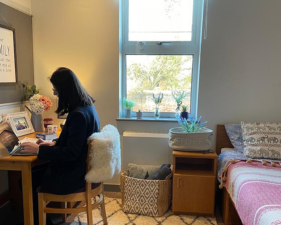 A girl sitting at a table writing on her computer with bed to the right