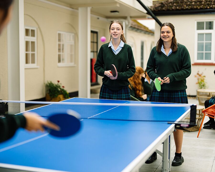 Two pupils in school uniform stand at a table tennis table holding paddles, while another person plays from the foreground.