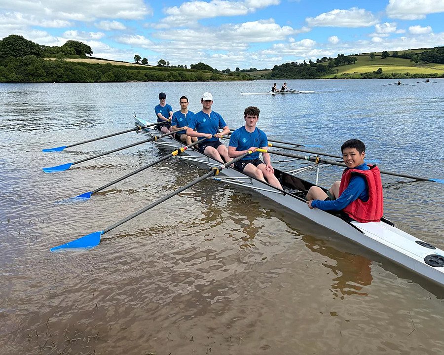 A rowing team of five students sitting in a boat on a calm lake surrounded by rolling hills.