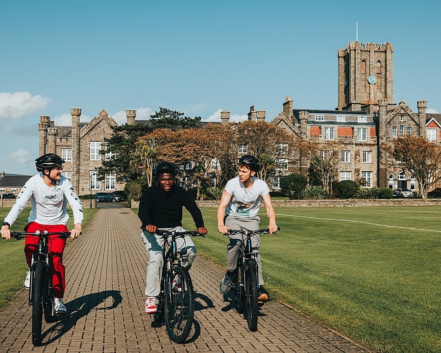 Three children on bikes cycling along path with school King Williams College in back
