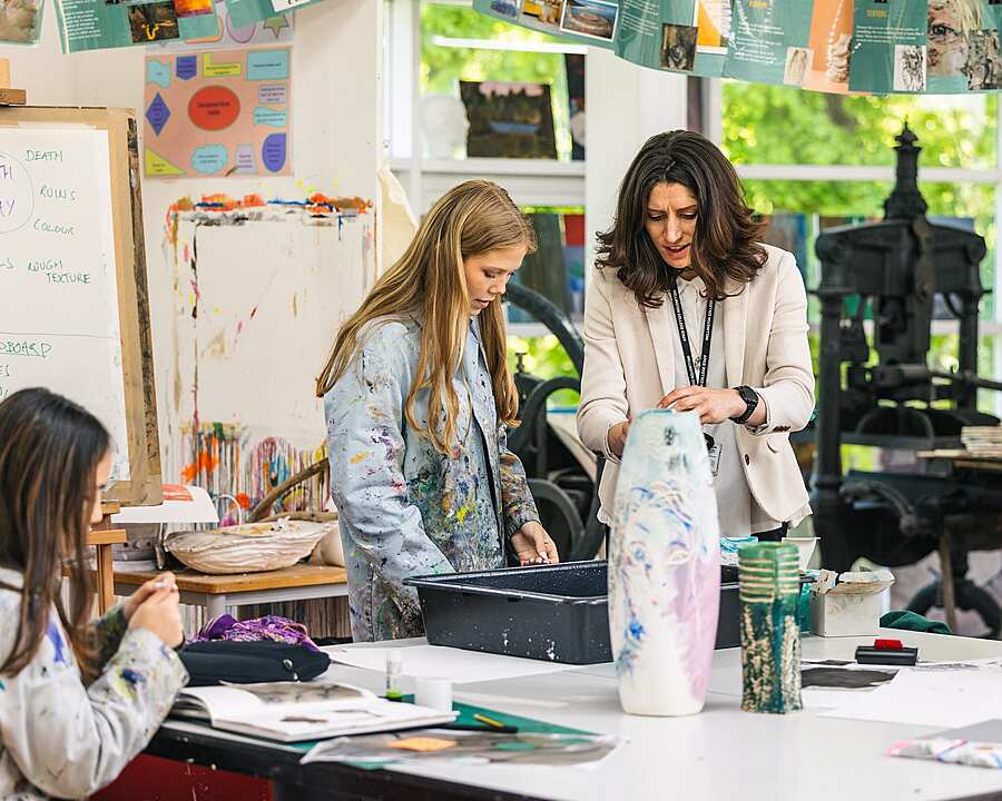 Teacher assisting a student with a ceramic art project in a bright classroom filled with creative materials.
