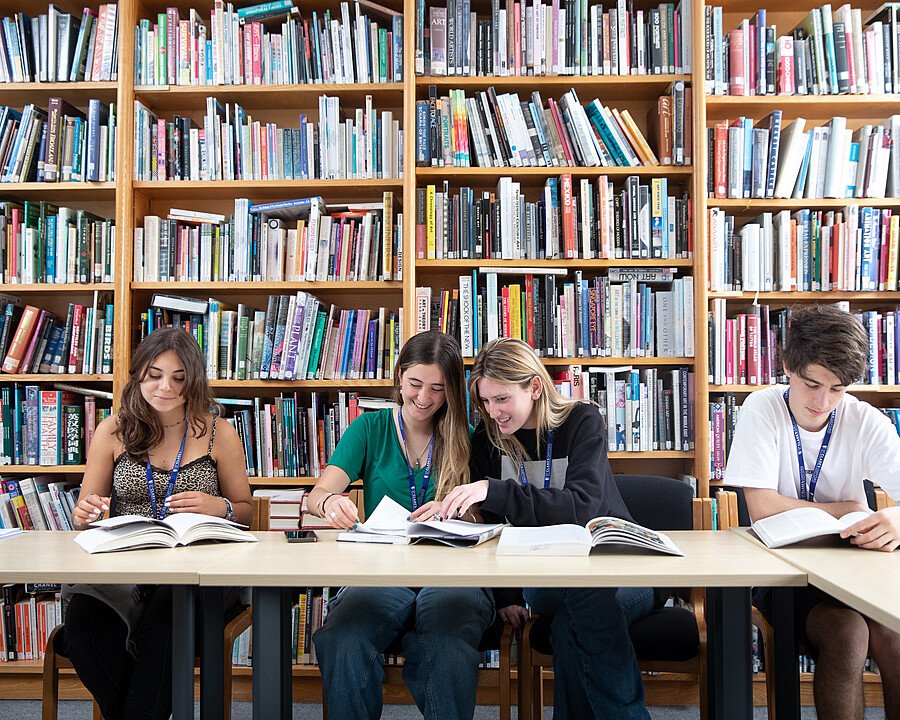 Four students studying together at a table in a library filled with bookshelves.