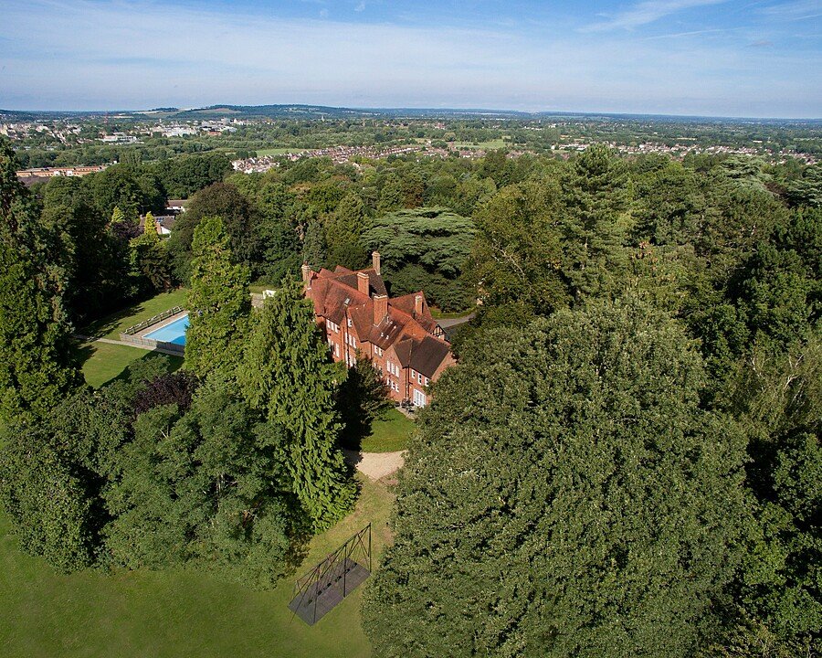 Aerial view of a large red-brick building surrounded by trees, with a swimming pool and expansive green grounds.