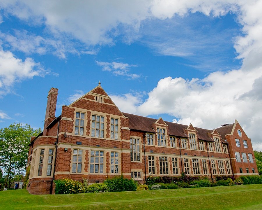 Large red-brick academic building with tall windows and manicured lawns under a partly cloudy sky.