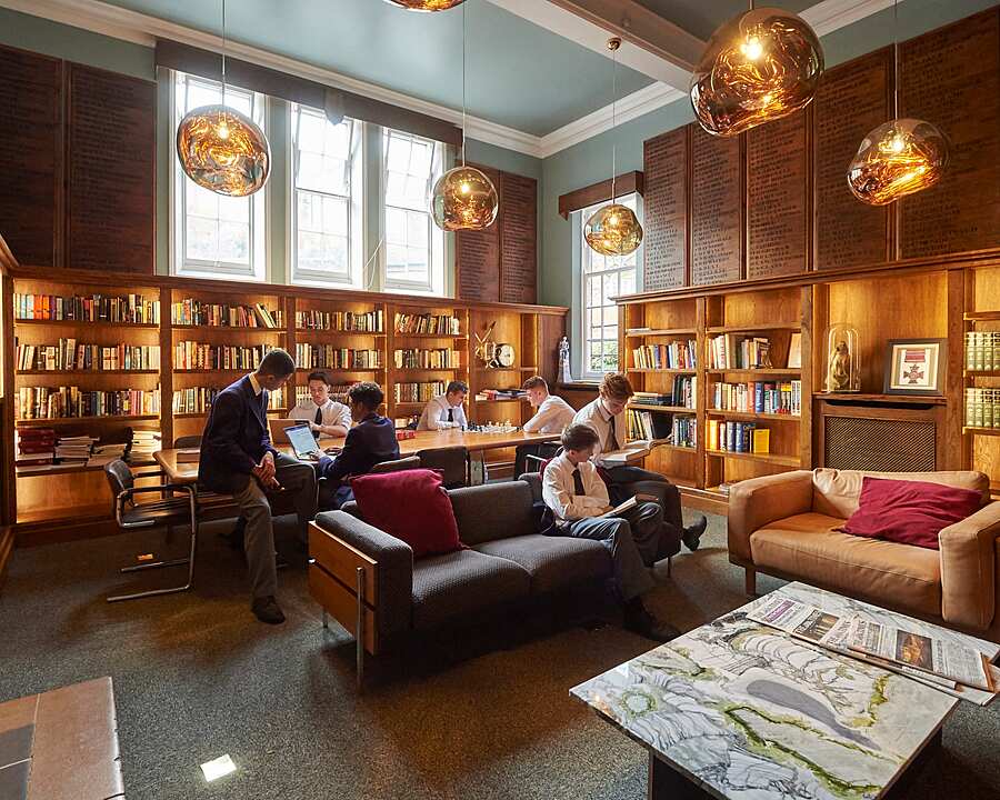 Students studying and reading in a cozy wood-paneled library with sofas and bookshelves.