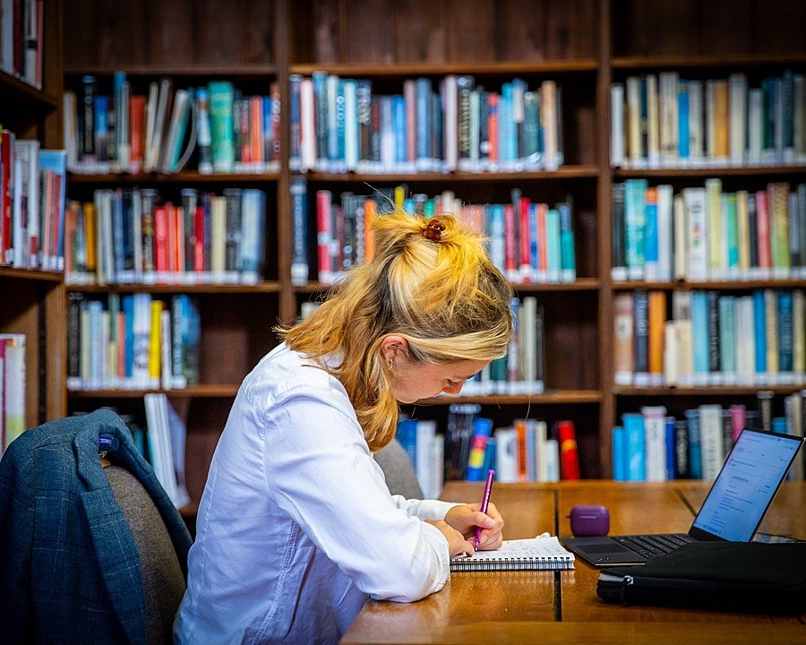 A student learning making notes on a notepad in front of an open laptop in a library