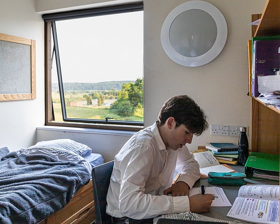 Boy sitting at a table in the process of writing with bed in the background