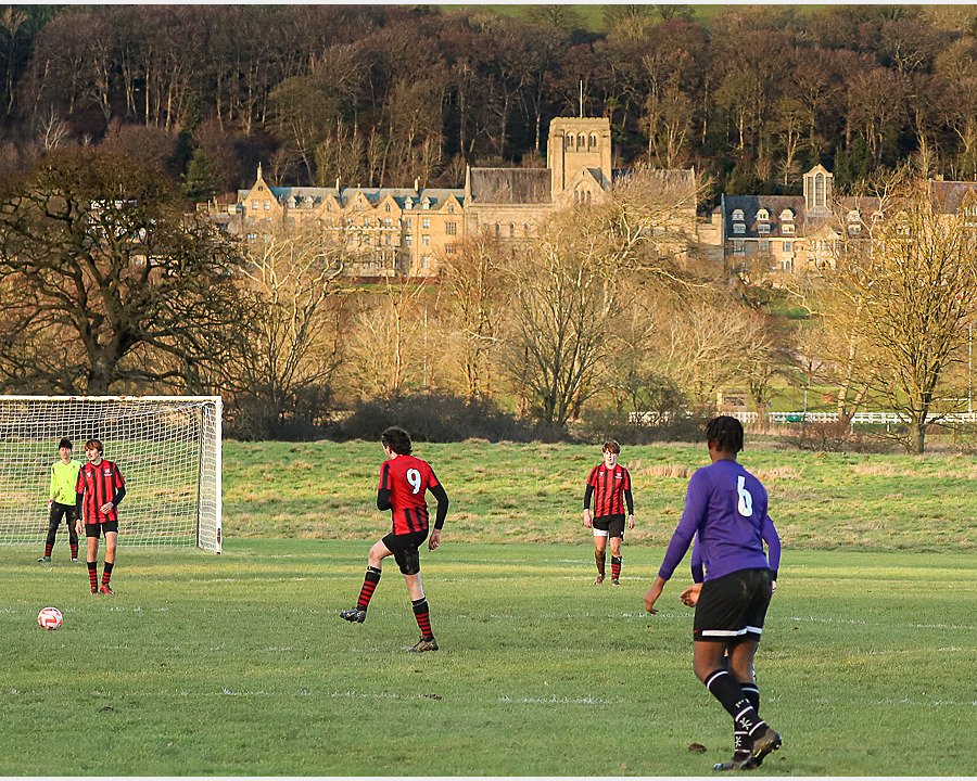 Boys in football jerseys playing a match outside, with a big building in the background