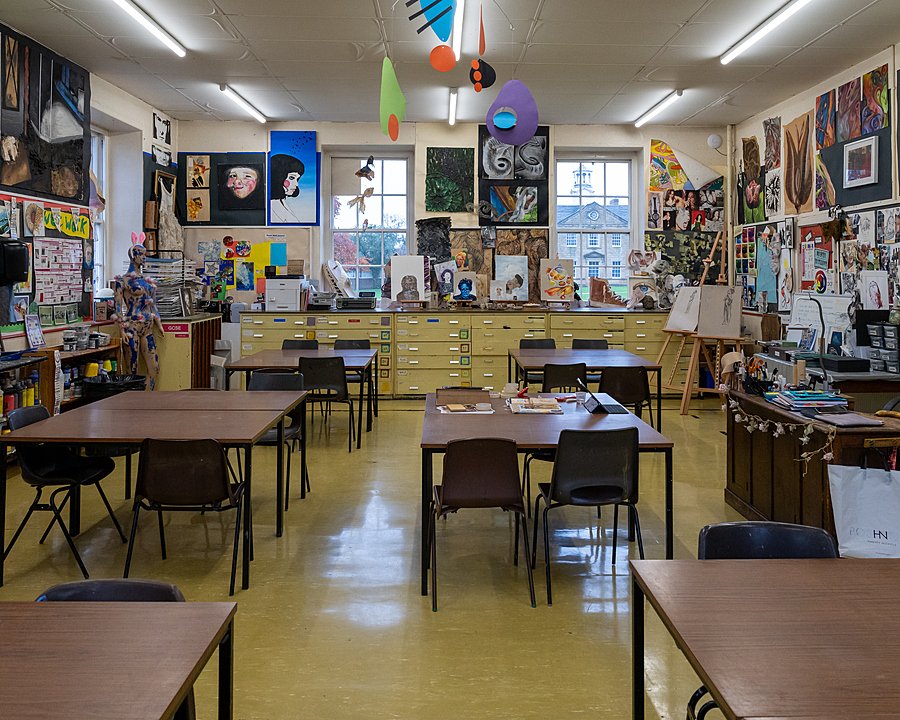 An art classroom filled with paintings, sketches, and creative projects covering the walls. Tables and chairs are neatly arranged, with art supplies and student work visible throughout the room.