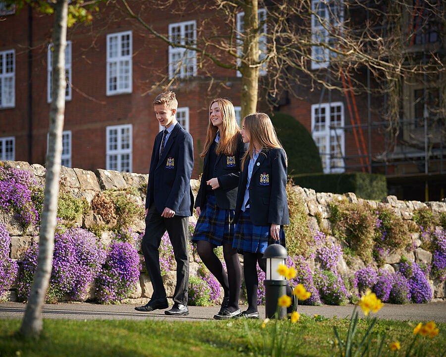 Three students in school uniforms walking beside a stone wall covered with purple flowers on a sunny day.
