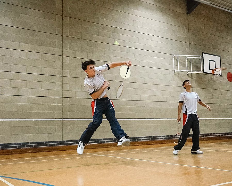 Two students playing badminton indoors, one jumping mid-air to hit the shuttlecock.