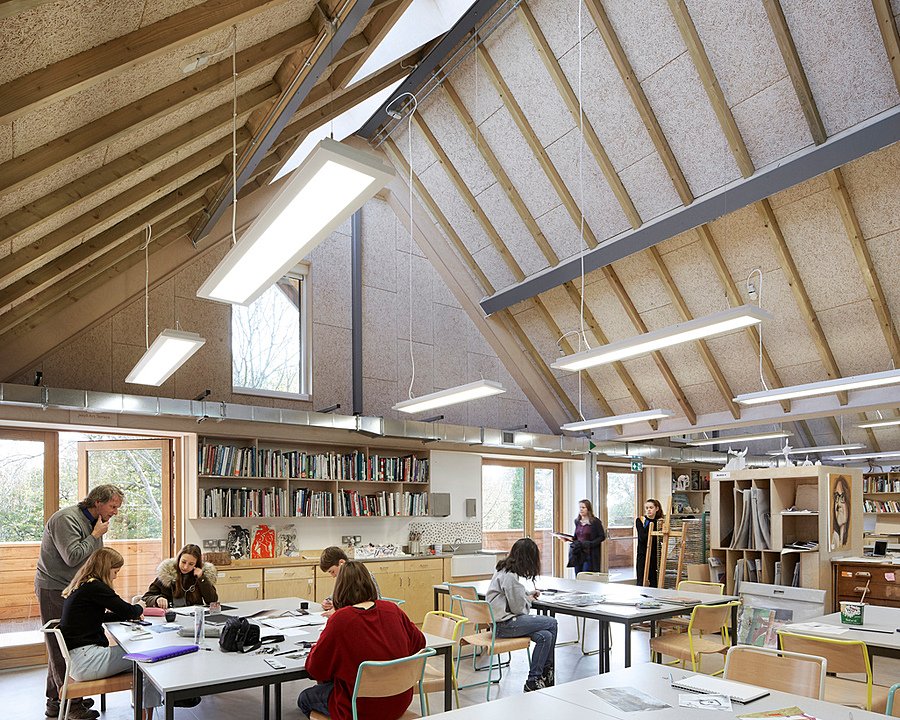 A bright art classroom with a high, beamed ceiling where students work on creative projects at large tables.