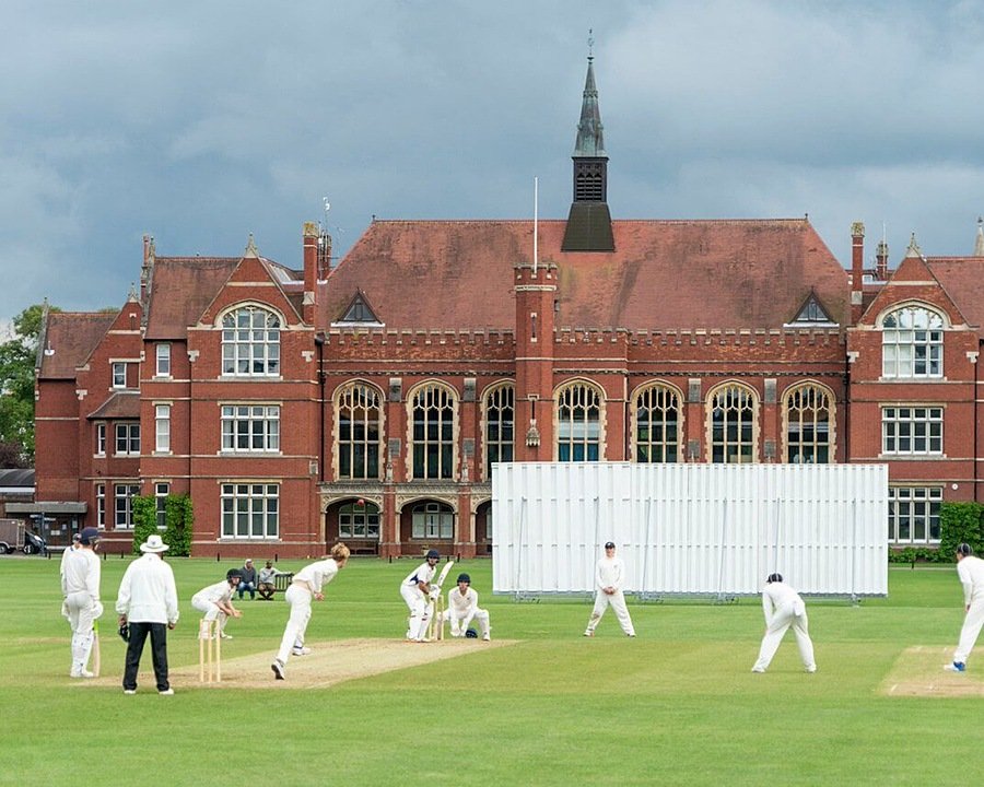 Boys in white cricket gear playing cricket with red-brick building as backdrop