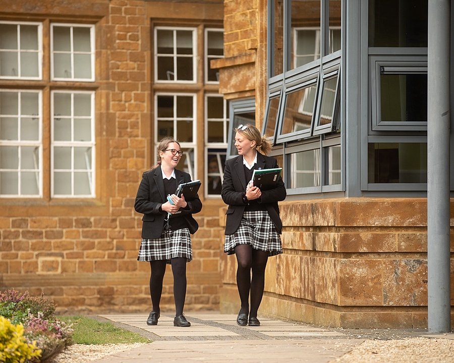 Two students in school uniforms walking and talking outside a stone school building.