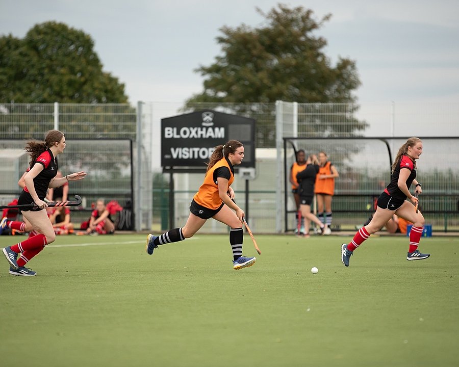 Three students playing field hockey on an outdoor pitch, with a “Bloxham Visitors” sign in the background.