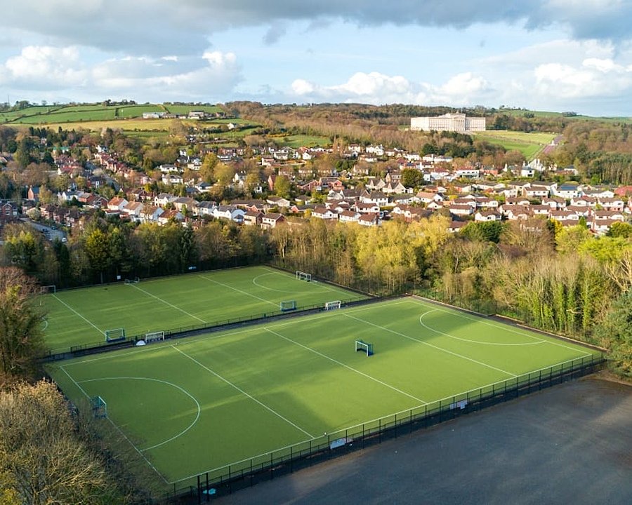 Aerial view of two green astroturf hockey pitches surrounded by trees and a nearby town.