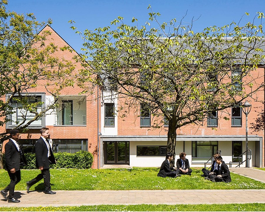 Students walking and sitting on the grass outside a modern red-brick school building on a sunny day.