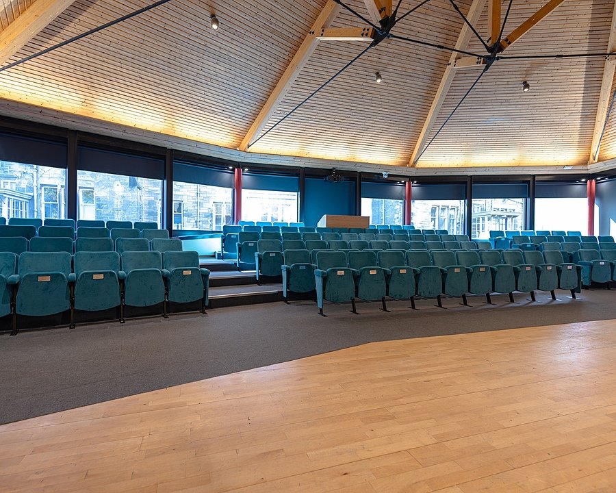 Modern circular lecture theatre with blue upholstered seats and wooden ceiling beams.