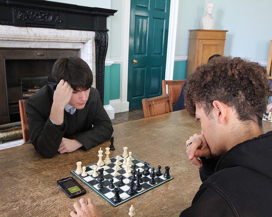 Two students deeply focused as they play a game of chess in a traditional study room with a fireplace.