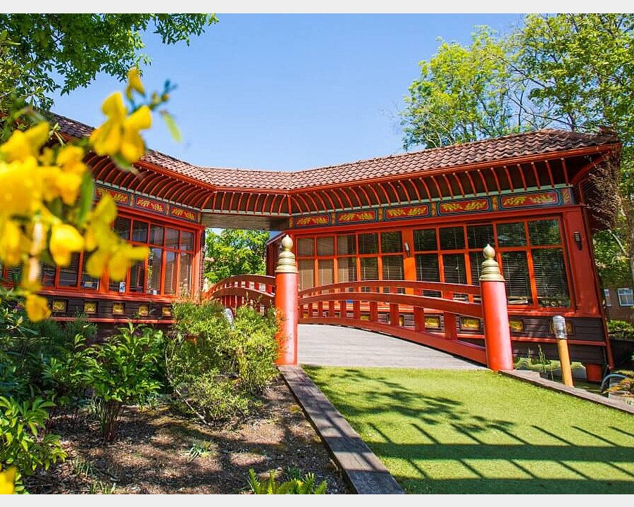 Decorative red pavilion with an arched bridge surrounded by greenery on a sunny day.