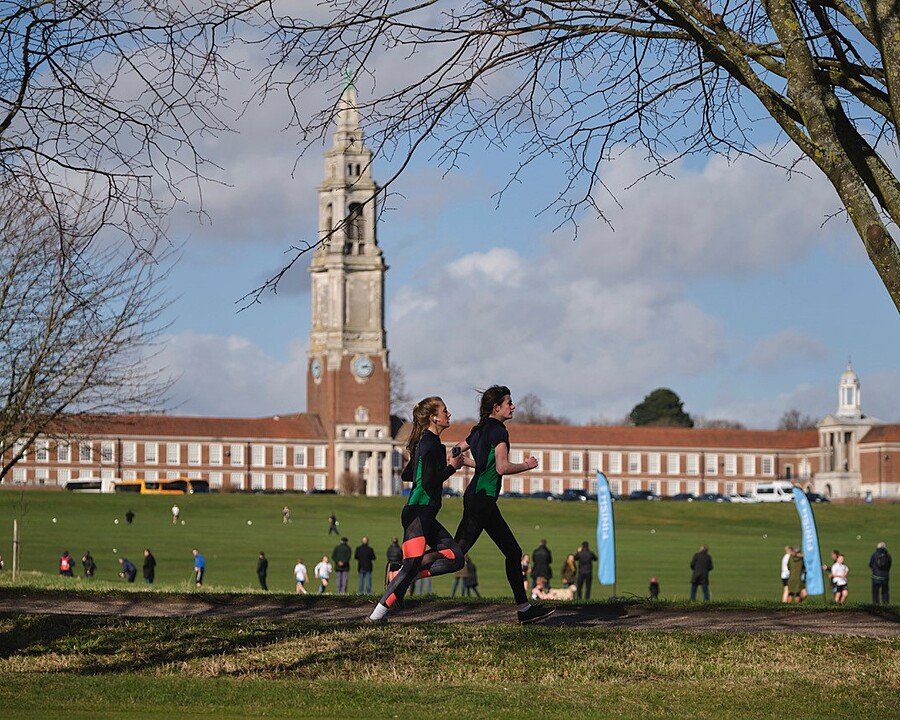 Two pupils running across a large grassy field with a grand school building and tall clock tower in the background under a partly cloudy sky.