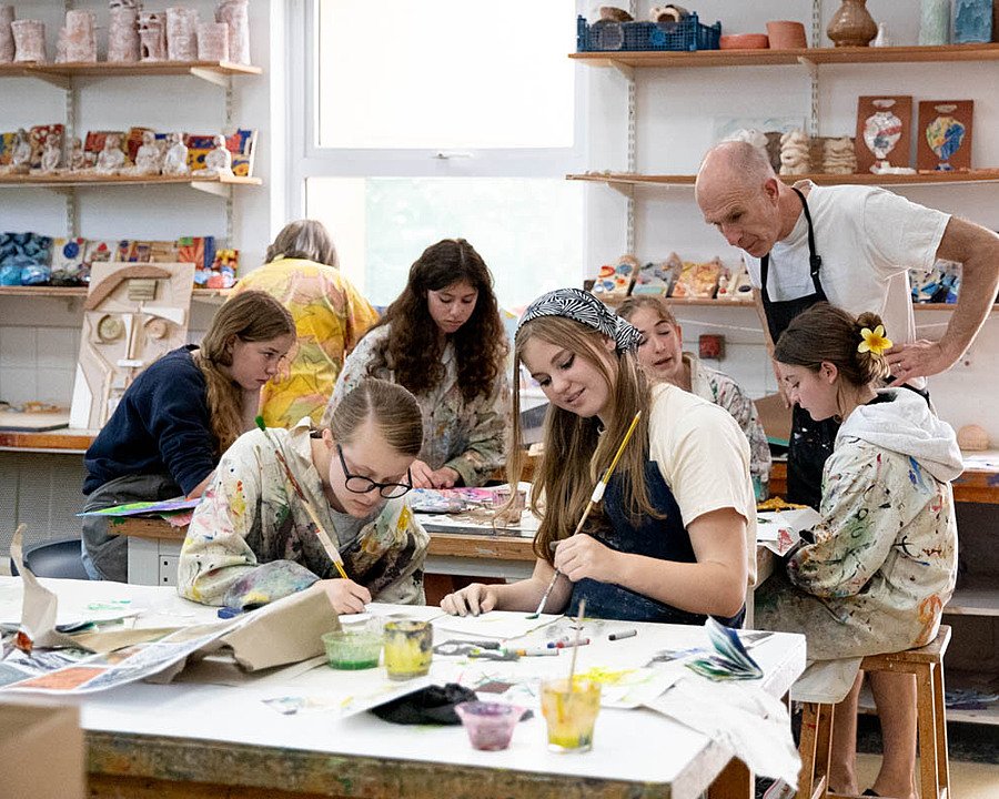 Art classroom where students in paint-splattered aprons are working on colourful projects under teacher guidance.