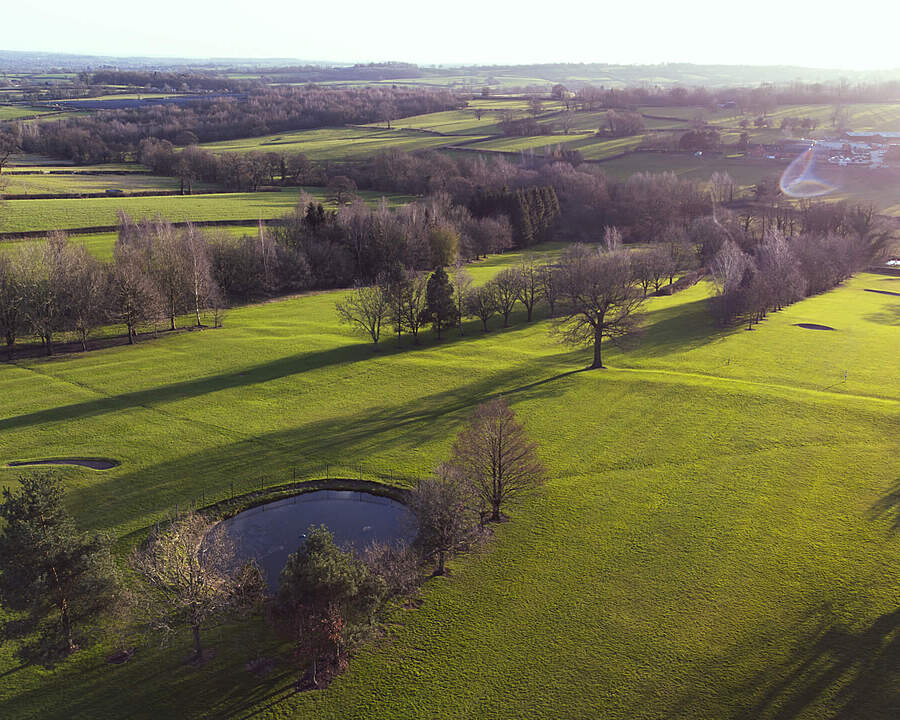 A scenic aerial view of a green landscape with open fields, scattered trees, and small ponds under soft sunlight.