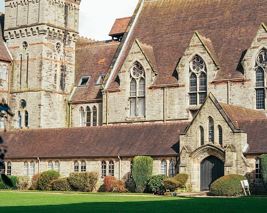 A large, historic stone building with arched windows and a tall tower, surrounded by neatly trimmed bushes and a green lawn.