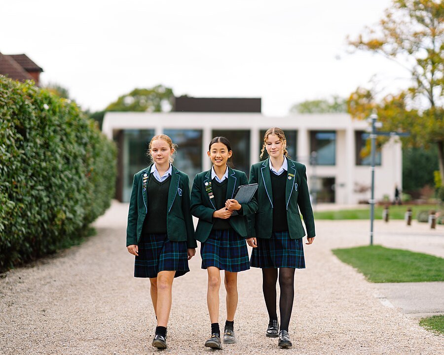 Three pupils in green school uniforms walk side by side along a gravel path on campus.