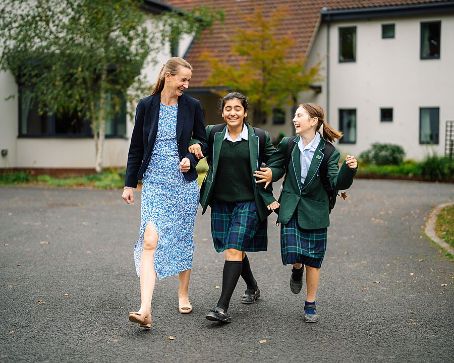 An adult woman walks with two pupils in school uniform along a paved path on campus.