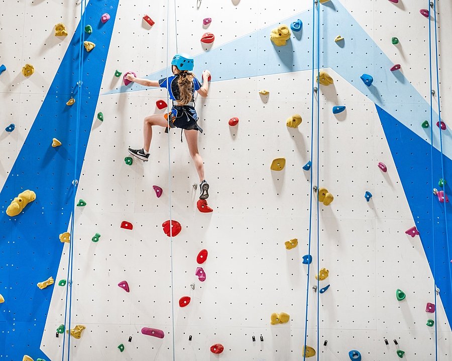 A climbing wall with a girl climbing up it, wearing a safety helmet