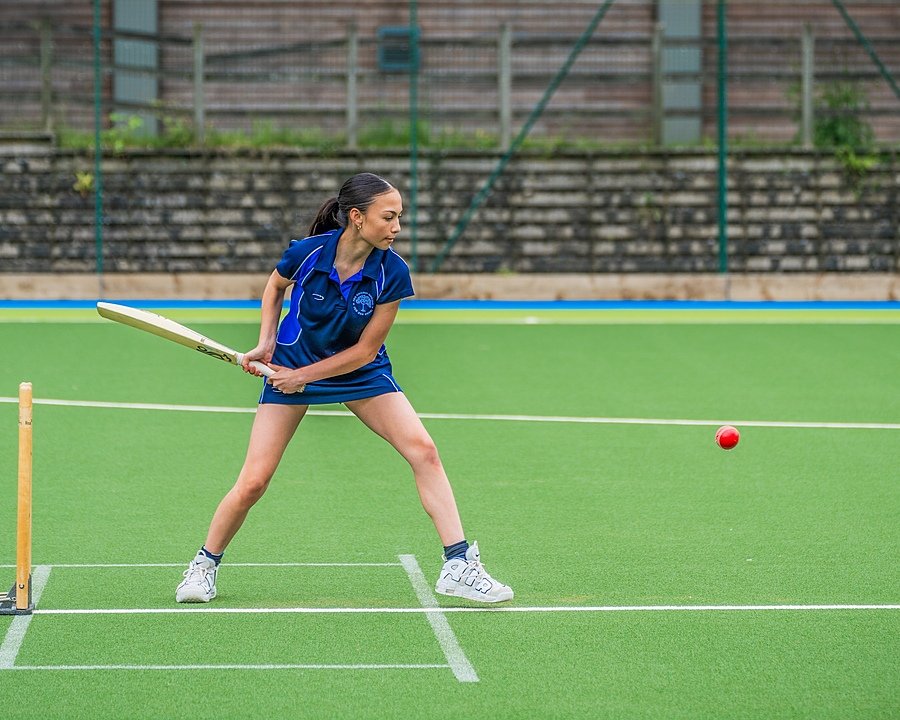 Girl in blue sports jersey holding a cricket bat, about to bat a red ball