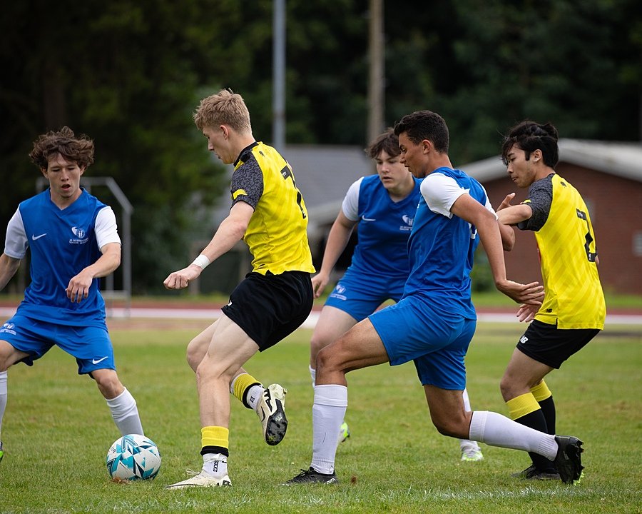 A group of students compete in a football match, with one team in blue and the other in yellow and black kits.