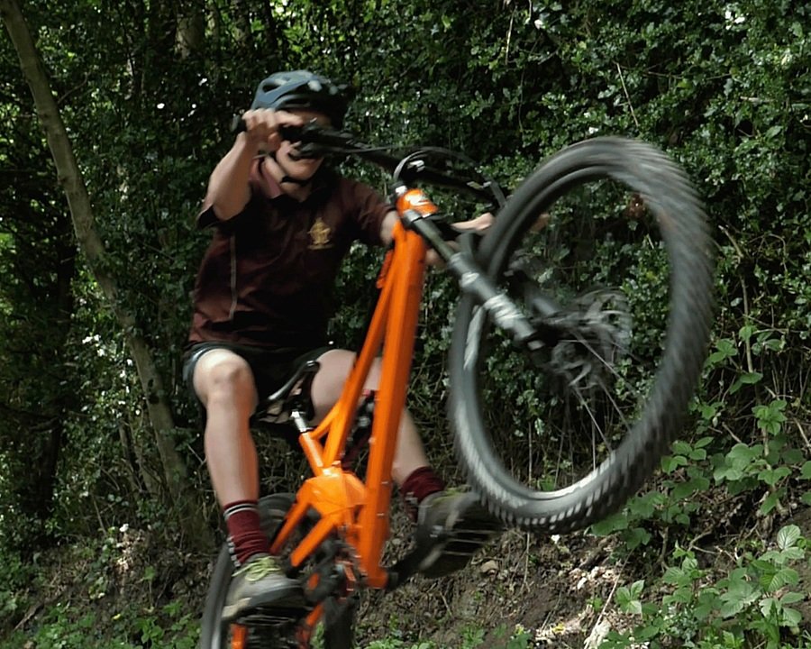 A student performing a wheelie on an orange mountain bike along a forest trail.