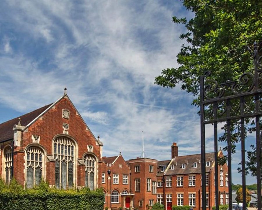 Traditional red-brick school buildings framed by wrought-iron gates and leafy trees under a blue sky.